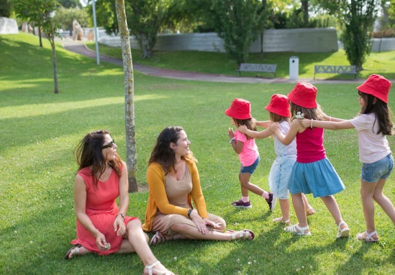 Happy children playing in a sunny park with two mothers sitting nearby, enjoying the outdoors.